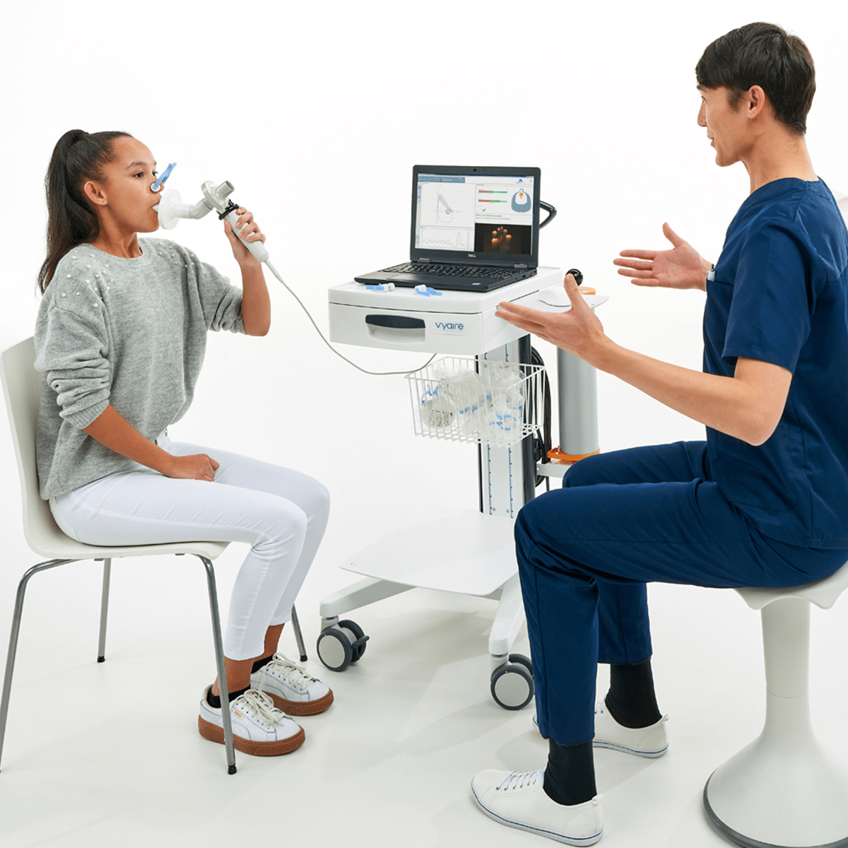 A girl does a breathing test with a spirometer while a healthcare professional monitors data on a laptop.