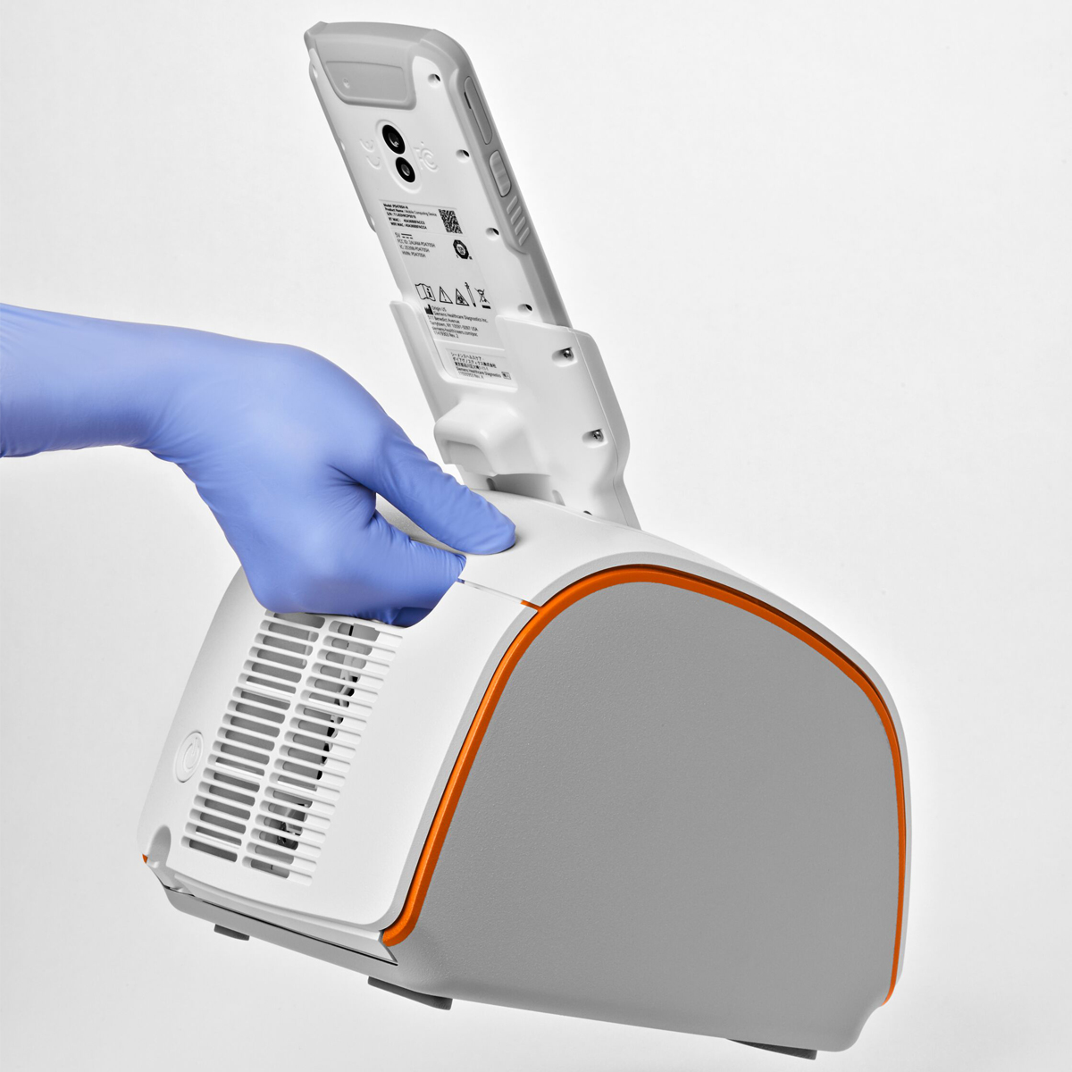 A gloved hand operates a white medical device with an orange trim and a handheld reader inserted on top.