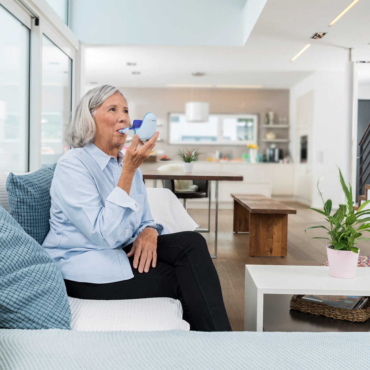 Older woman sitting on a couch using an inhaler device in a bright, modern living room.