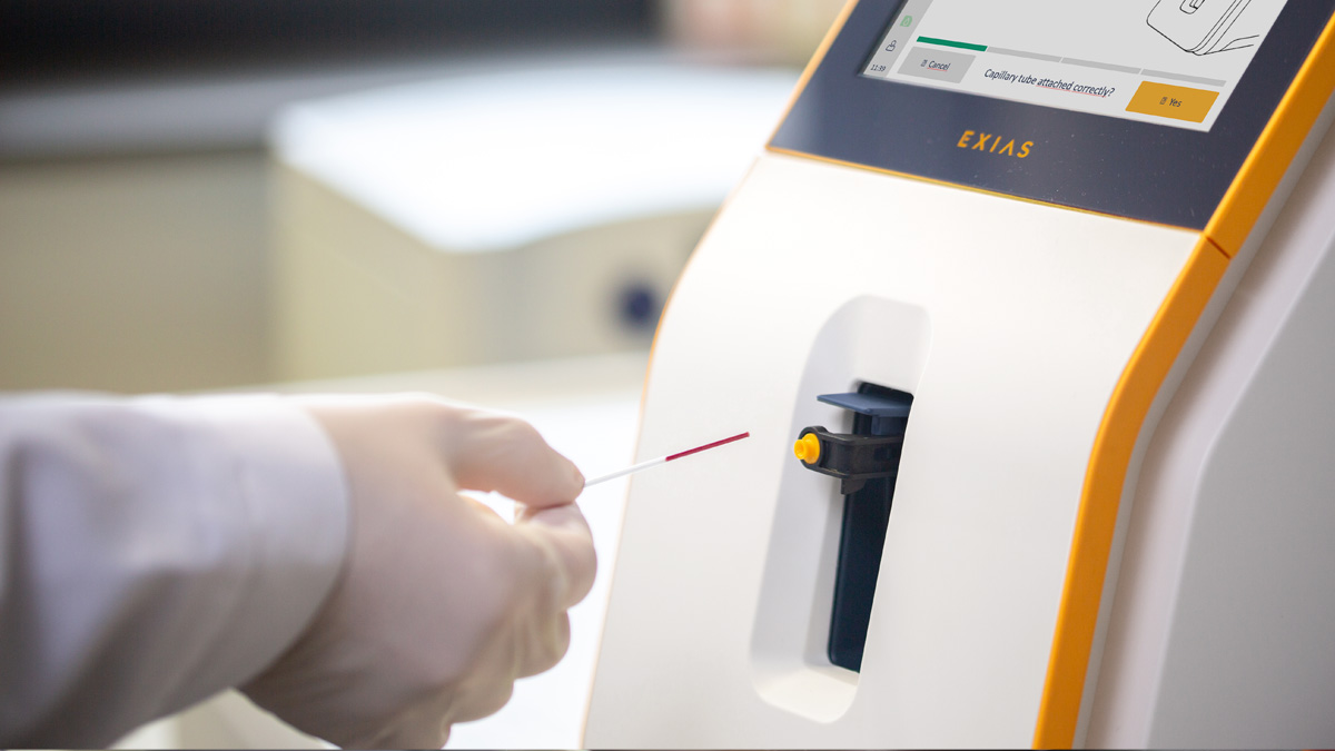 Gloved hand inserting a blood sample tube into a medical analyzer machine with a display screen.