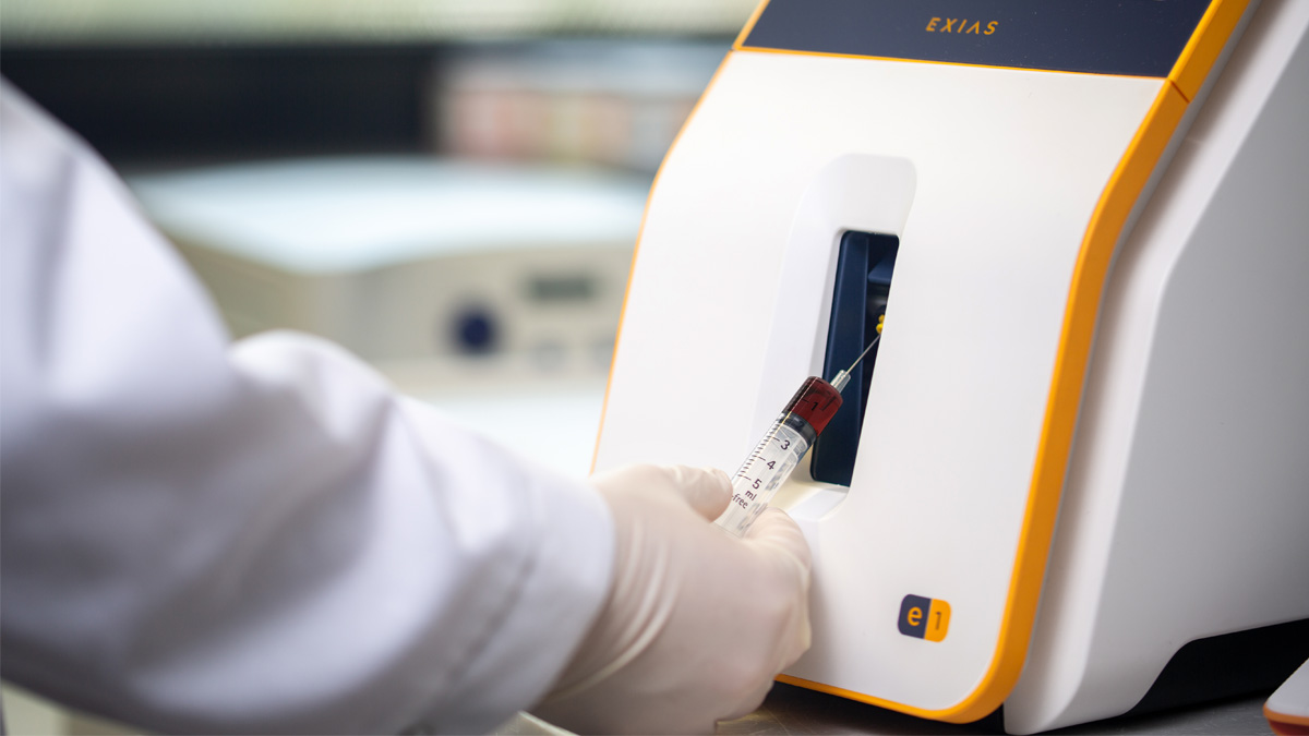 Gloved hand inserts a blood sample syringe into a medical analyzer machine labeled "EXIAS" for testing.