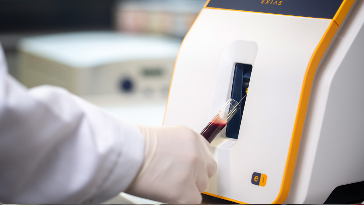 Gloved hand inserting a blood sample tube into a laboratory analyzer machine.