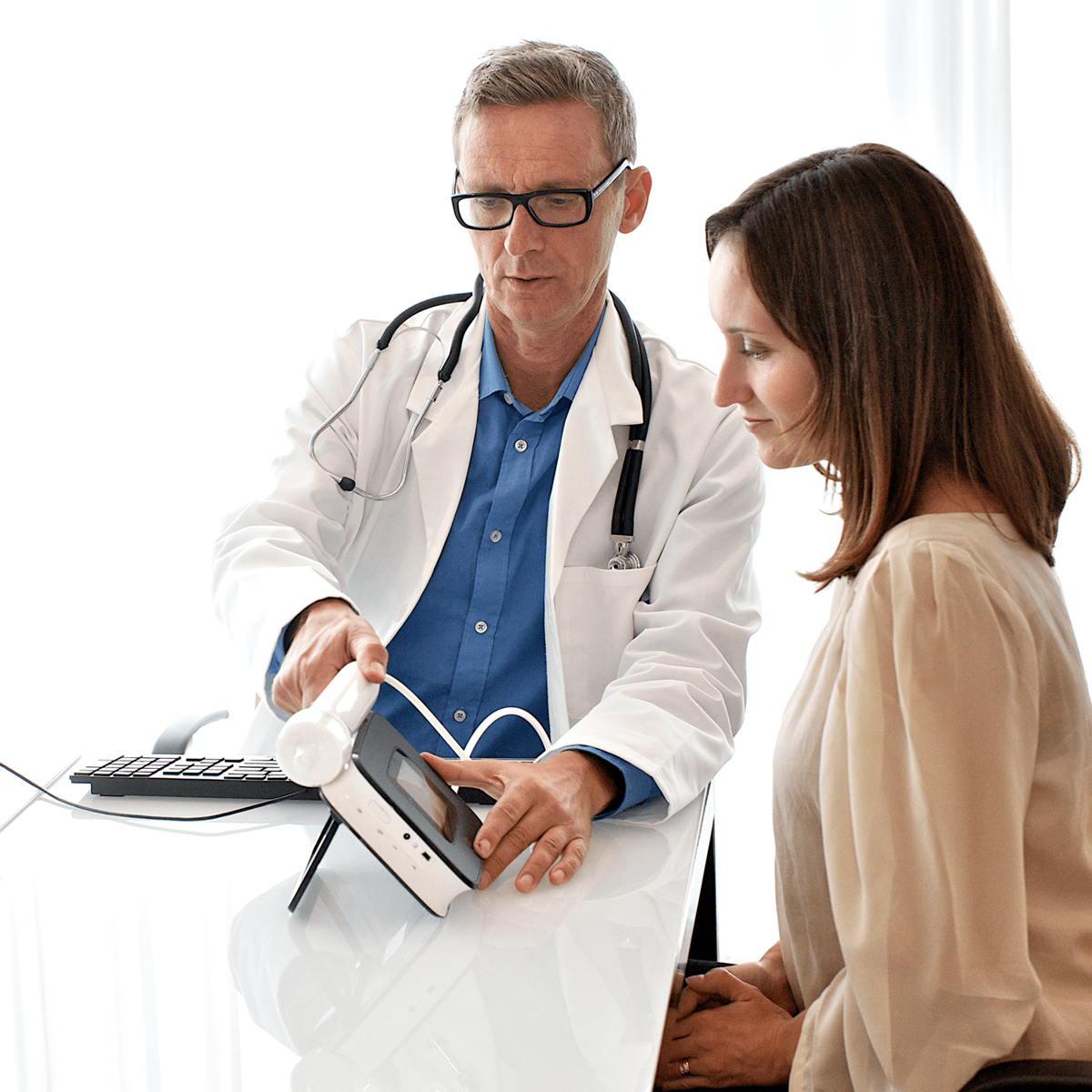 Doctor showing a female patient a medical device at a white desk in a bright office setting.