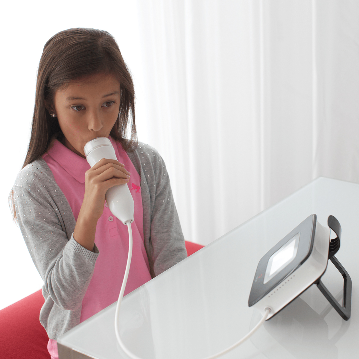 Girl using a spirometer, blowing into a medical device on a table for a lung function test.