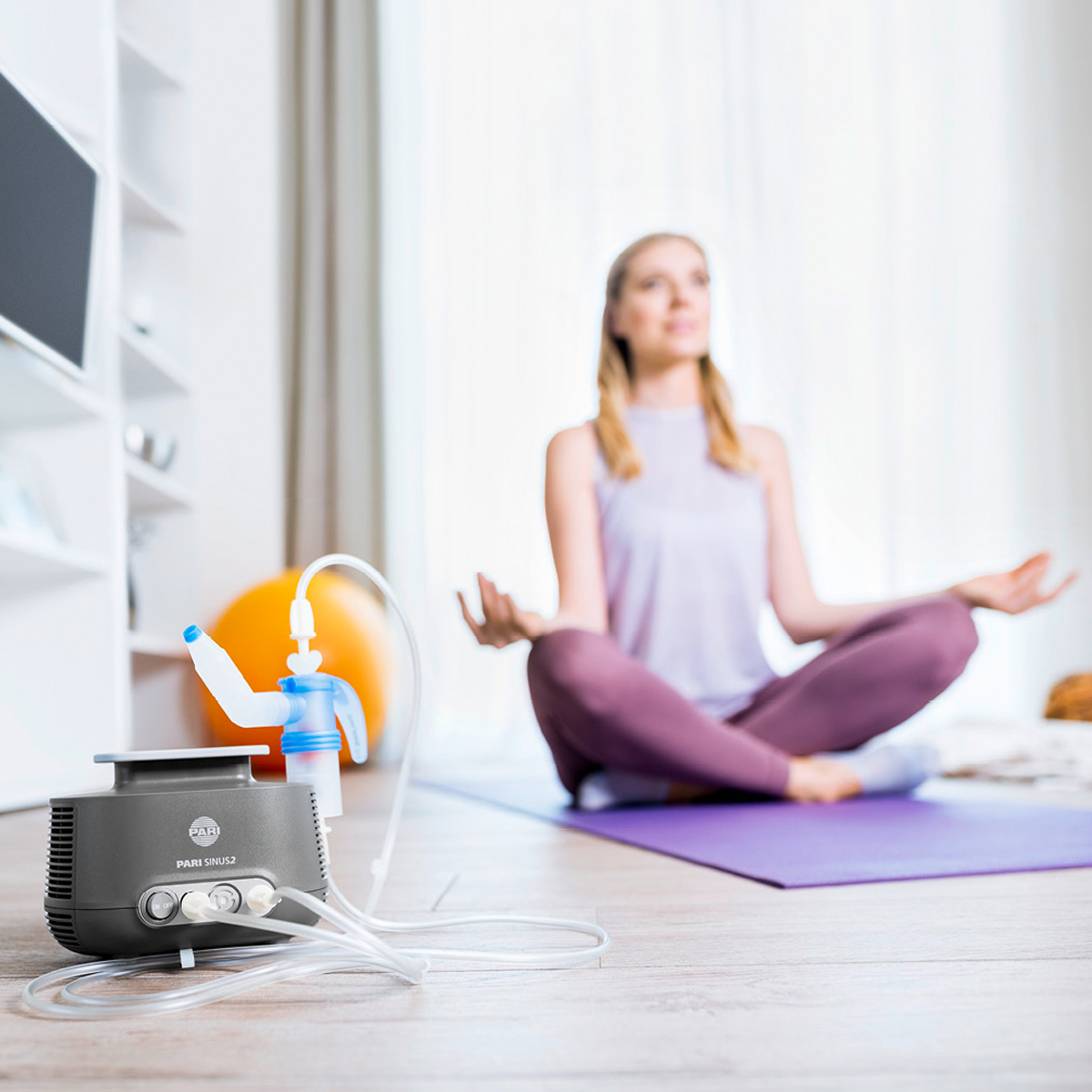 A woman sits cross-legged on a yoga mat, with a nebulizer machine in the foreground.