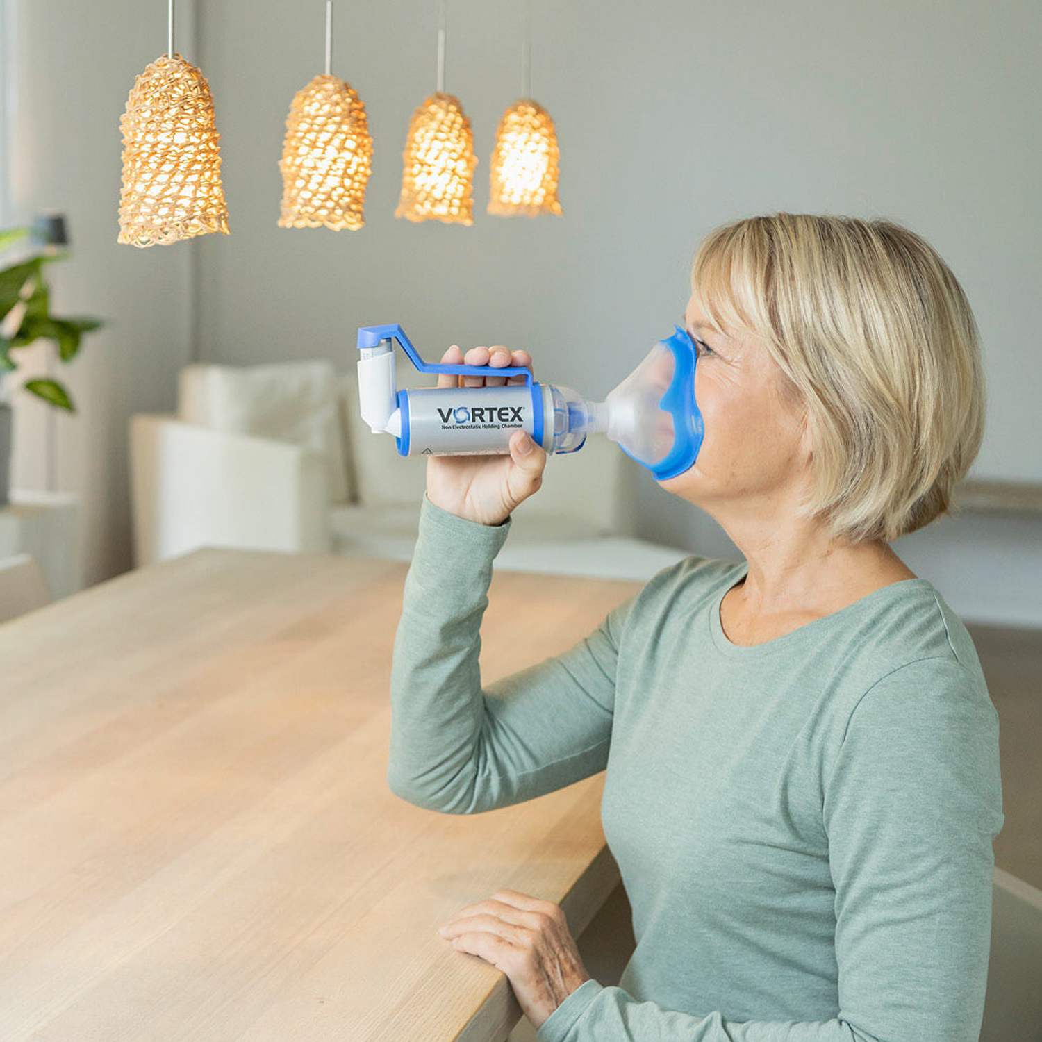 Woman using an inhaler with a spacer at a table in a well-lit, modern room with hanging lights.