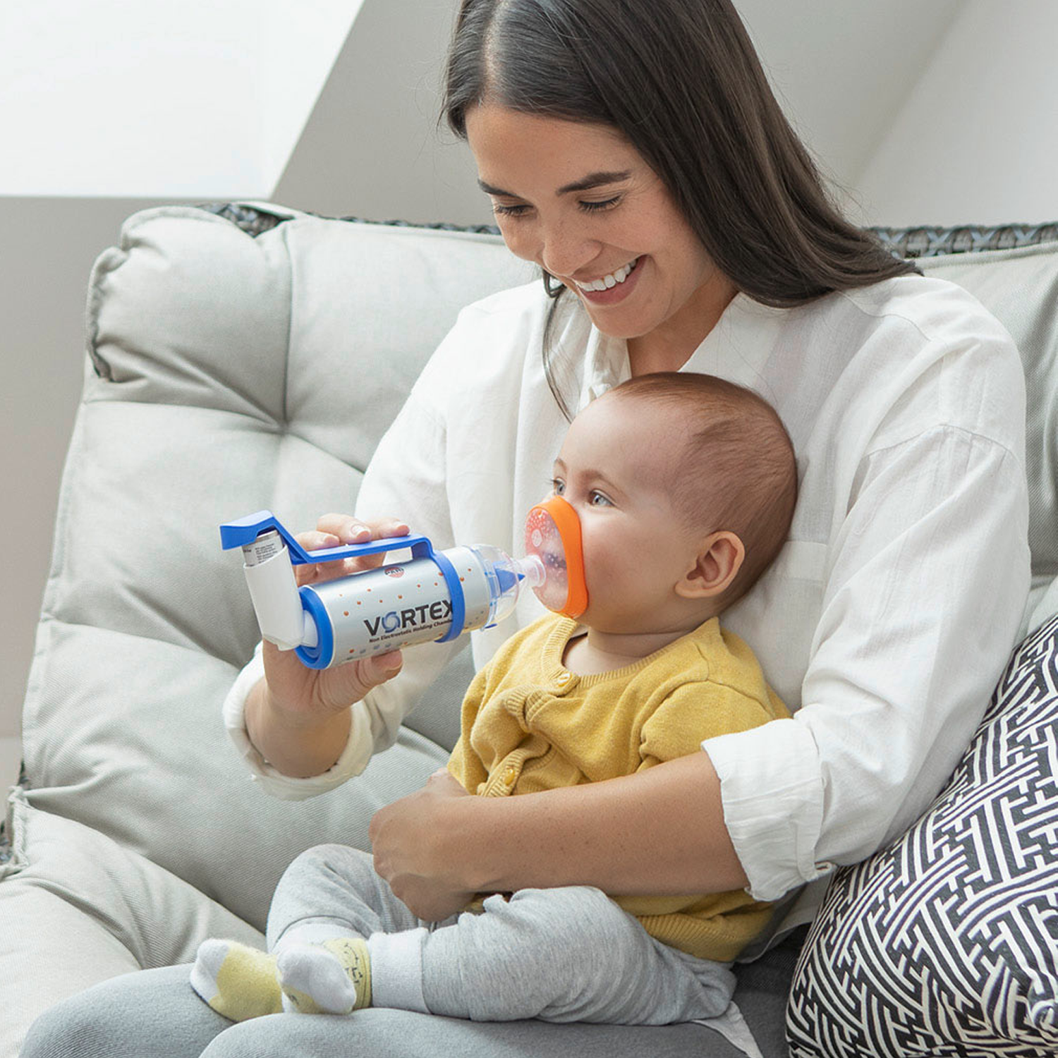 Woman smiling while giving a baby an inhaler with a mask, sitting on a couch.