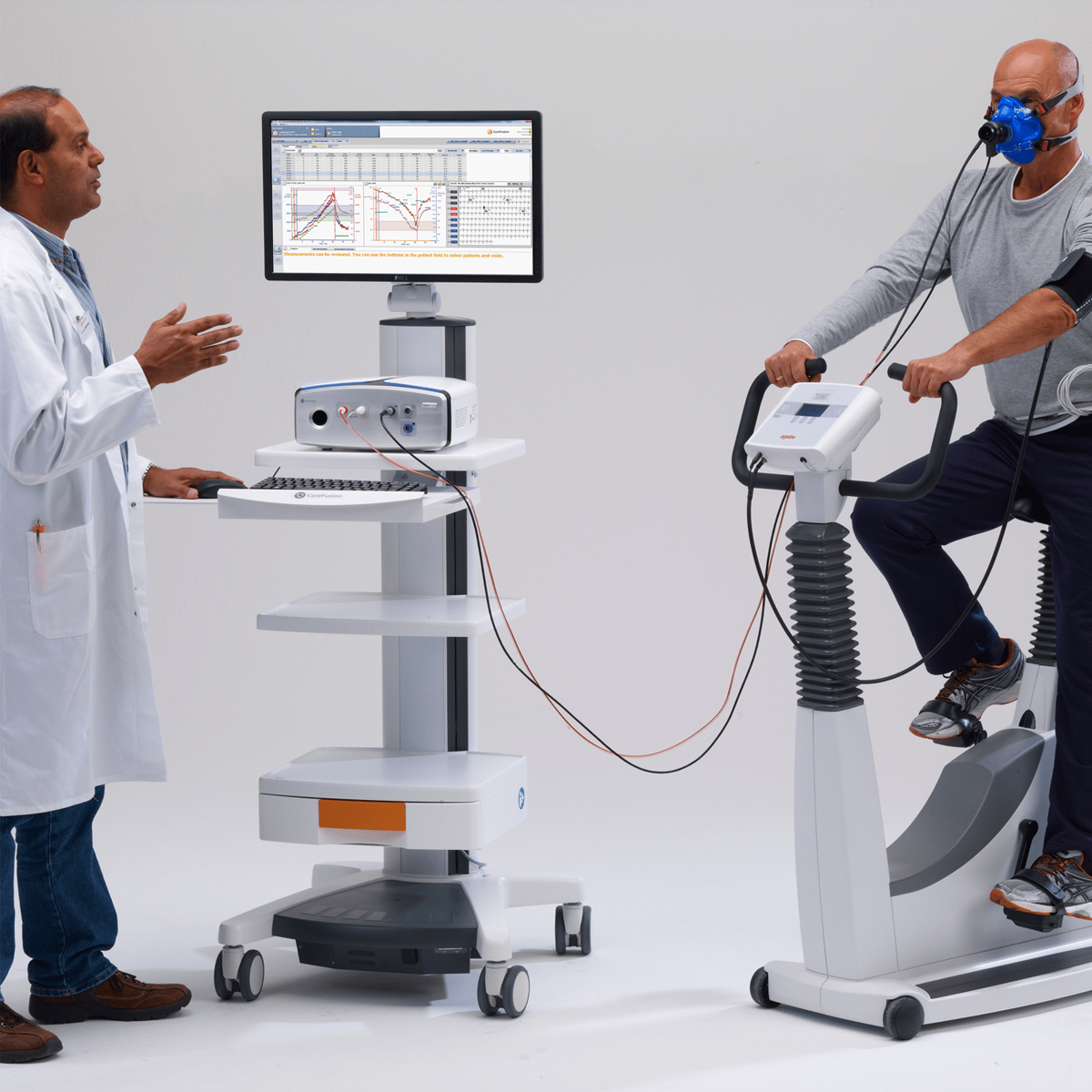 Doctor monitors a patient on an exercise bike wearing a breathing mask, with medical equipment and a chart on display.