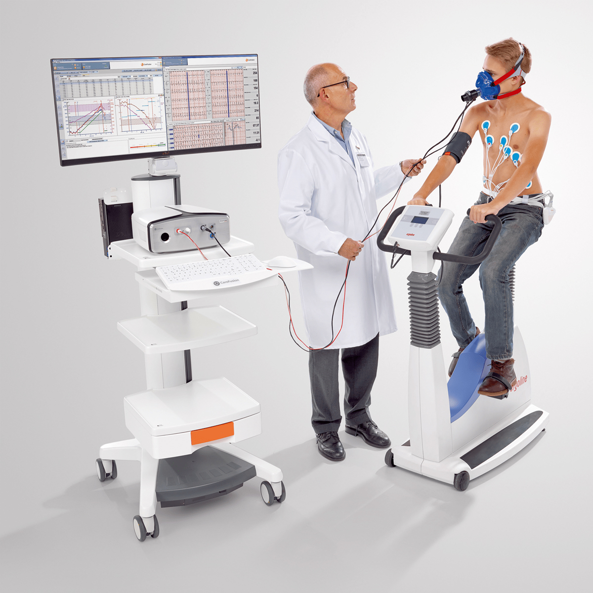 Doctor supervises a man on an exercise bike wearing sensors and a mask; medical equipment and monitor nearby.