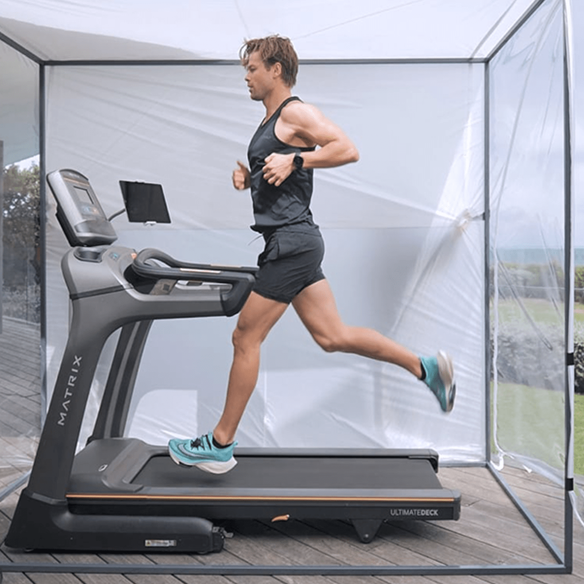 Man running on a treadmill indoors, wearing black athletic outfit and turquoise shoes.