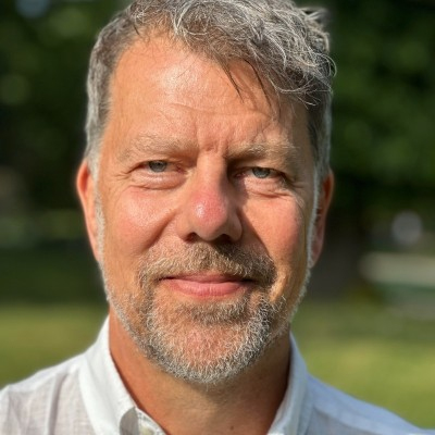 Middle-aged man with gray hair and beard smiling outdoors, wearing a white collared shirt, green background.