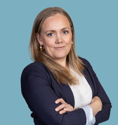 Woman with long blonde hair in a navy blazer and white shirt, arms crossed, against a blue background.