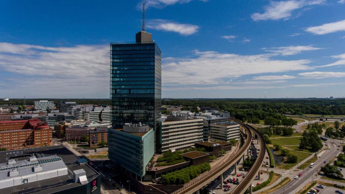 Tall glass office building surrounded by smaller buildings, train tracks, and green landscape under a blue sky.