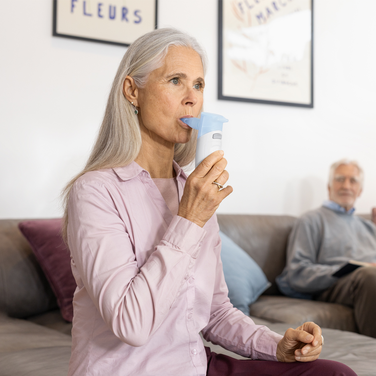 Older woman using an inhaler or nebulizer while sitting on a couch, with an older man in the background.