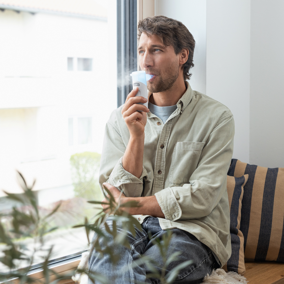 Man sitting by a window using an inhaler, wearing a light-colored shirt, with plants in the foreground.