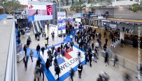 Busy convention center entrance with people walking on a large "WELCOME" floor sign and digital screens above.